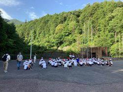 青々とした山に囲まれた広場で、子どもたちが地べたに座って正面に立つ2名の生徒の方を見ている写真