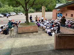 駐車場と建物の間の広いスペースで、関係者の方々が周りで立っているなか、子どもたちがきれいに並んで座っている写真