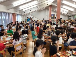 ごはんが用意されたテーブル席がずらりと並んだ部屋で、子どもたちが席に座り、両手を合わせていただきますの仕草をしている写真