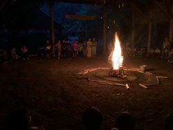 暗くなった屋外で、キャンプファイヤーの火が勢いよく立ち昇り、あたりをほのかに照らしている写真