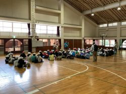 リュックを背負った子どもたちが、体育館に集まって正面に立つ関係者の方を向いている写真