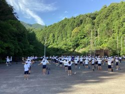 青々とした山に囲まれた広場で、きれいに整列した子どもたちが体操をしている様子の写真