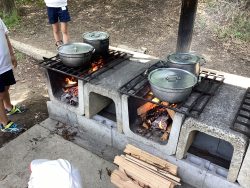屋外に備え付けられた複数のかまどに火がつけられ、それぞれのかまどに2つずつの大鍋が乗り湯気が立っている写真