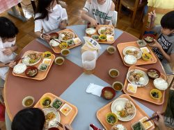 テーブル上に、ごはんや汁物、おかずが配膳されたトレーが7名分用意され、子どもたちがそれぞれお箸を手に食事をしている写真
