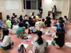 ピンク色のカーペットが敷かれた部屋にたくさんの子どもたちが集まって座っており、壁側に立っている2名の生徒が紙を見ながら話をしている様子の写真