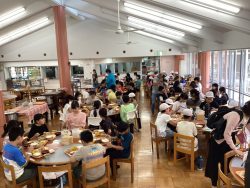 丸いテーブル席がずらりと並んでいる部屋で、関係者が所々に立って子どもたちを見て回っている様子と、椅子に座った子どもたちが食事をしている写真