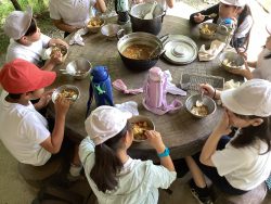 2つの大きい鍋が中央に置かれた丸いテーブルを、子供たちが囲んで座っており、一緒にカレーを食べている写真