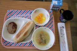 縞模様のランチマットを敷いたトレーに、食器に入ったパンやおかず、スープやデザートが配膳され、紙パックの牛乳やコップと一緒にテーブルに並んでいる写真