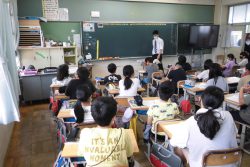 椅子に着席している子どもたちと、黒板の前に立っているワイシャツ姿の先生の写真