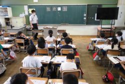 ひらがなの文字が書かれた黒板と、椅子に座っている子どもたち、黒板の横に立っている先生の写真
