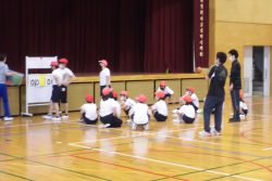 体育館に体操服姿の子どもたちが座っており、3名の生徒がスコアボードの周囲に集まっている写真