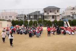 校庭にたくさんの子どもたちが整列して座っており、左側の列の方に数名の子どもたちが駆けつけている様子の写真