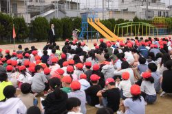 校庭に座っているたくさんの子どもたちと、スーツ姿で立って子どもたちの方を向いている先生の写真