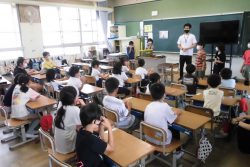 黒板の前に、男性の先生と数名の子どもが横並びに立っており、椅子に着席している子どもたちの方を見ている写真