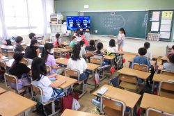 モニターに画像が映し出された教室で、数名のこどもたちが黒板の前に立って、着席している子どもたちの方を向いている写真