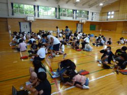 体育館内でたくさんの子どもたちがタブレットを操作しており、関係者の人たちが見回りながら子どもたちの様子を見ている写真