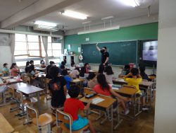椅子に着席している子どもたちと、黒板の前に立って右手を上げている黒い服を着た先生の写真