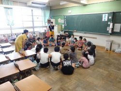 机を端に寄せて広い空間を作った教室で、子どもたちが輪を作って床に座っている様子の写真