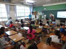 モニターが用意された教室で、モニターの横に立って話をしている関係者と、椅子に着席して関係者の方を見ている子どもたちの写真