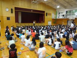 子どもたちが体育館に座って集まっており、正面に立つ先生の方を見ている写真