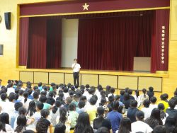 たくさんの子どもたちが、舞台下に立って話をしている先生の方を向いて座っている写真
