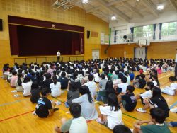 体育座りをしている子どもたちが体育館に集まっており、正面に立つ先生の方を見ている写真