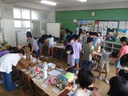 教室内の机をくっつけて並べた上に、様々な作品が展示されており、たくさんの子どもたちが作品を見て回っている様子の写真