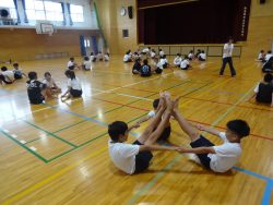 体育館内に、三人一組になった子どもたちが散らばって座っており、手前側の3人は両手両足を繋げるようなポーズをとっている写真