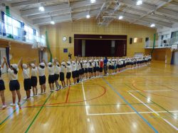 体育館を斜めに区切るように横一列になった子どもたちが両手を上に挙げてポーズをとっている写真