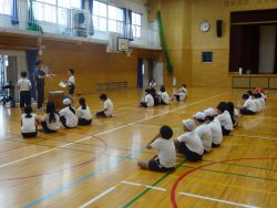 体育館の床に座っている子どもたちと、子どもたちの方を向いて手振りを加えながら話をしている先生の写真