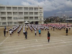 校庭にずらりと並んだ子どもたちと先生方が立っている様子を後ろから撮った写真