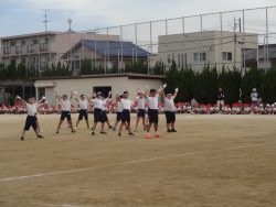 周囲からたくさんの子どもたちが座って見守る中、校庭の中央あたりに立つ子どもたちが両手をあげてポーズをとっている写真