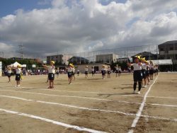 校庭に間隔をあけて並び、黄色の帽子姿で両手を前の方に出している子どもたちの写真