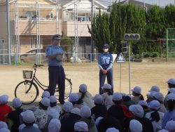 自転車や小さい信号機が用意された校庭で、白い帽子を被って座っている子どもたちの正面に、2名の警察官の方が立って話をしている様子の写真