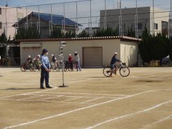 校庭で、小さな信号機の横に警察官の方が立っており、校庭の奥の方には自転車にまたがっている関係者が待機している様子の写真