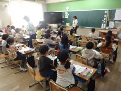 机に紙や筆記道具を置いてイスに着席している子どもたちと、教室内を歩いて回る関係者たちの写真
