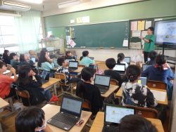 モニターの横に立って話をしている先生と、開いたタブレットを机の上に置いて先生の方を見ている子どもたちの写真