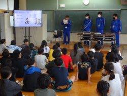 教室の正面に青のジャージを着た4名の中学生が並んでおり、左側に立つ中学生がモニターを使って話をしている様子と、床に座って話を聞いている子どもたちの写真