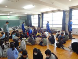 教室の床に座って話し合いをしている様子の子どもたちと、教室内を歩いて回る中学生や関係者の写真