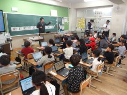 教室内に3名の関係者が立っており、椅子に着席している子どもたちがタブレットを開いた状態で正面の方を見ている写真