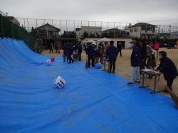 傾斜が付くように設置されたビニールシートが校庭に用意され、参加者や関係者の人たちが大きなサイコロを使ってイベントを行っている様子の写真