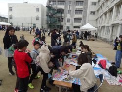 校庭におもちゃが置かれた机が用意され、参加者の人たちが列を作って机の前に並んでいる写真