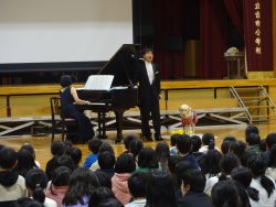 体育館の舞台下にグランドピアノが設置され、ドレス姿の山鹿さんが演奏をする中、スーツ姿の濱田さんが横に立って歌っている様子の写真