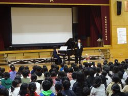 グランドピアノを弾いている山鹿さんと、歌っている濱田さんの様子を、床に座って鑑賞しているたくさんの子どもたちの写真