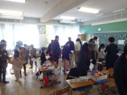 机や椅子を両端に寄せた教室内で、保護者の人と一緒に列を作って並んでいる子どもたちの写真
