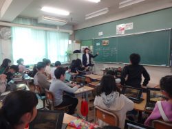 黒板を指さしている先生と、前列の子どもが席から立ち上がって先生の方を向いている写真