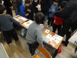 カードの様な紙が広げられている机と、保護者と子どもで賑わっている教室内の写真