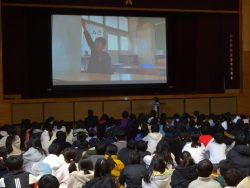 体育館で、設置されたスクリーン上に右手を挙げた1名の子どもが映し出されており、そのスクリーンを座って見ている子どもたちの写真