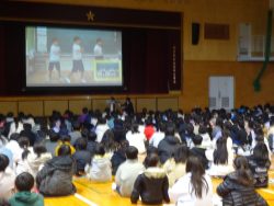 体育館で、設置されたスクリーン上に体操をしているような3名の子どもが映し出されており、そのスクリーンを座りながら見ている子どもたちの写真