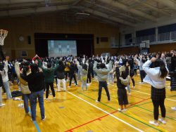 体育館に集まったたくさんの生徒たちが、スクリーンの方を見ながら両手を上げて体操をしている様子の写真
